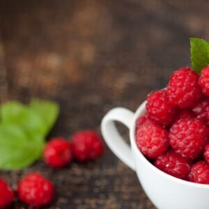 A close-up of juicy raspberries in a white cup with a green leaf accent on a rustic wooden table.
